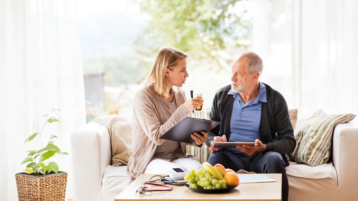 Das Bild zeigt die Situation einer Pflegebegutachtung. Eine Begutachterin spricht dafür mit einem Senior auf einem Sofa
